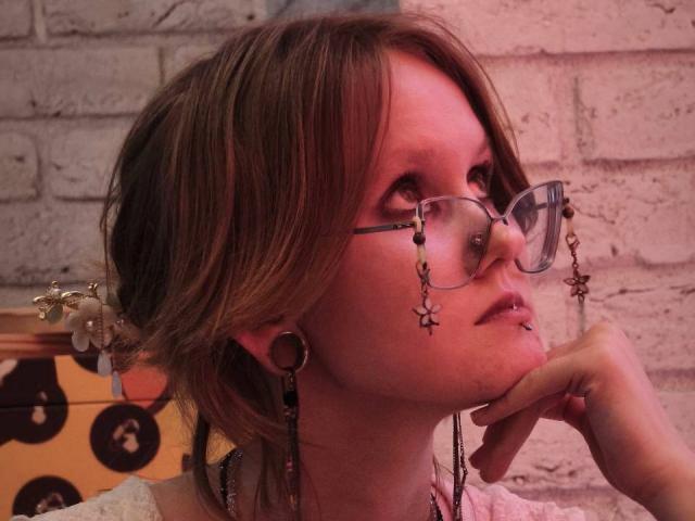 MozellContos has short brown hair, wears glasses with star-shaped pendants, has a nose ring, and a lip piercing. She's looking up with her hand on her chin.