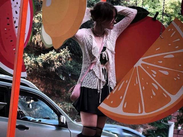 MozellContos with short brown hair, wearing lace pink top, black skirt, and thigh-high stockings, holding orange slices, standing by a silver car, pink purse slung over shoulder.