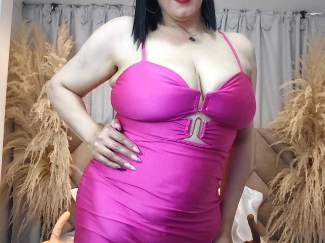 Liza Russell mit großen, runden Brüsten in einem engen, pinken Kleid, eine Hand am Hüfte, schwarzes Haar, roter Lippenstift und einer kleinen Anhängerkette, steht vor beige farbenen Vorhängen..