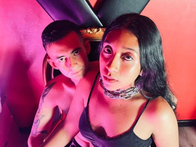 LiaMarek with black hair, wearing a black tank top and silver chain, sits next to a shirtless LiaMarek with a tattoo on his right arm. Pink lighting.