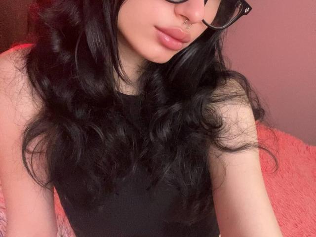 GlitchDoll com longos cabelos pretos ondulados, blusa preta sem mangas, piercing no nariz e óculos pretos, mostrando seus lábios rosados e curvados, pele clara e sua bunda quase imperceptível no enquadramento..