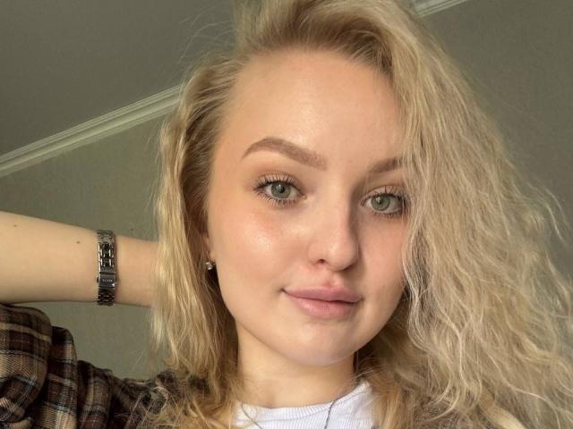 NievesMegee has blonde wavy hair, green eyes, and a slight smile. She's wearing a plaid shirt and a silver bracelet. Her face is slightly shiny.