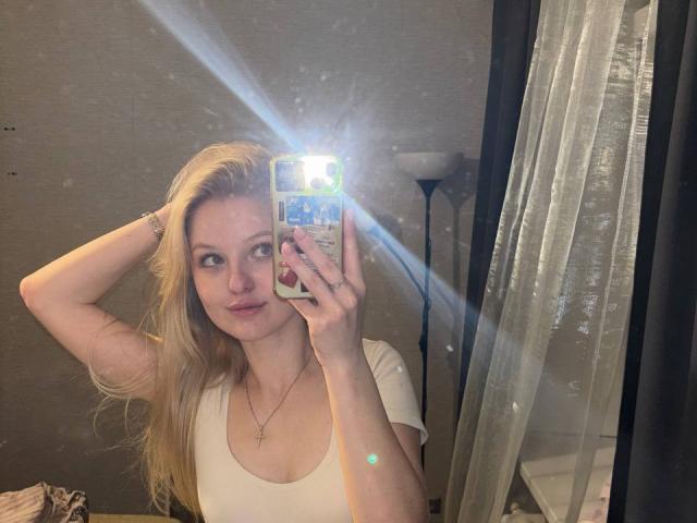 NievesMegee with blonde hair, white tee, holding phone, flash in mirror, hand in hair, small cross necklace, subtle smile, visible reflection, standing in dimly lit room.