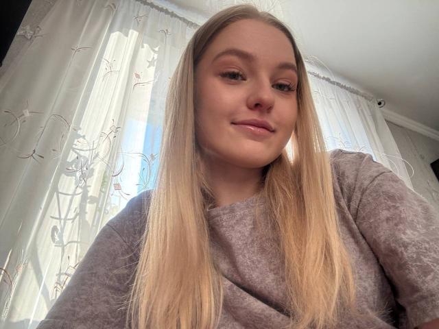 NievesMegee with long blonde hair, wearing a grey sweater, has a subtle smile, light makeup, and a slightly flushed face, looking down at the camera.