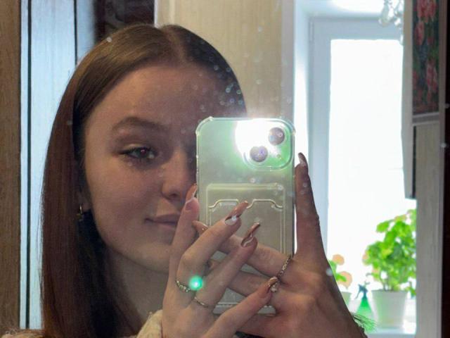NievesMegee with long brown hair, holding a phone with a glowing ring, taking a selfie in a mirror, showing a subtle smile, and wearing a ring on her finger.