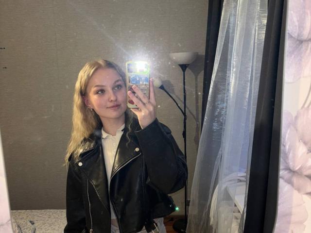 NievesMegee with blonde hair, wearing a black leather jacket, white shirt, taking a mirror selfie, holding a phone with a blue and white case, light reflection in mirror.