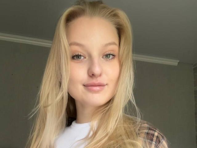 NievesMegee has blonde hair, green eyes, and a slight smile, wearing a white shirt with a plaid jacket, showing her cute face and small, perky ass.