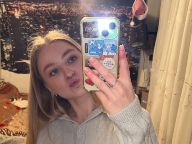 NievesMegee with blonde hair, pouty lips, holding phone, wearing gray hoodie, showing ass-like phone case, ring on finger, mirror selfie, soft light, casual bedroom.