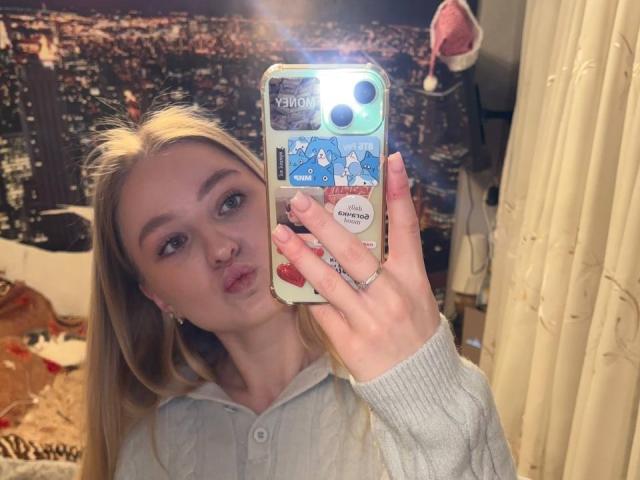 NievesMegee with blonde hair, pouty lips, and light makeup, holding a phone with a blue cartoon sticker case, wearing a grey hoodie, in a mirror selfie.