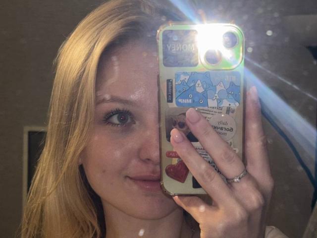 NievesMegee with blonde hair, holding a phone with a map case, pouting her pink lips, and looking into a mirror with a bright flash. Her ring is visible.