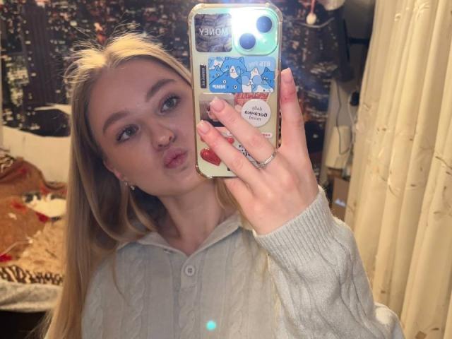 NievesMegee with blonde hair, pouty lips, wearing a gray rib-knit hoodie, taking a mirror selfie with a phone featuring a sticker of cute blue animals.