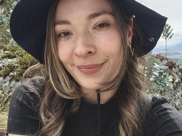 TaniaSandersX, con un sorriso carino, occhi marroni e capelli castano chiaro ondulati, indossa un cappello nero e una maglietta nera, mostrando il naso e le labbra leggermente dischiuse..