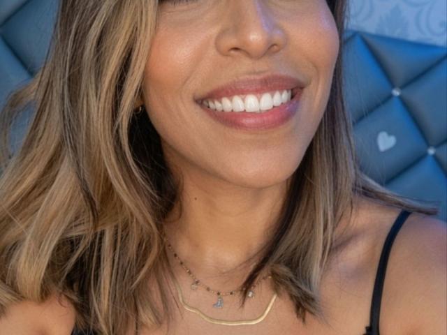 AuroraBenson's smiling face, showing white teeth and pink lips, with blonde hair, wearing a black strap top and gold necklace, featuring small blue gems.