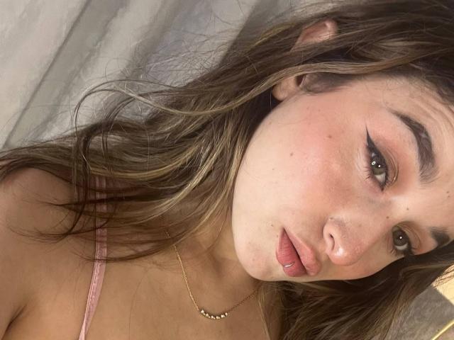 MilaGonz has pouty pink lips, a gold necklace, and brown wavy hair. She's wearing a pink bra, with winged eyeliner and a slight blush on her cheeks.