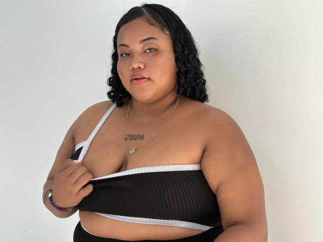 Alisha Monroe, eine übergewichtige Frau mit lockigem, schwarzem Haar, hält ihren schwarzen und weißen BH hoch und zeigt ihre kleinen Brüste und ein Tattoo über ihrer Brust. Sie trägt eine Halskette..