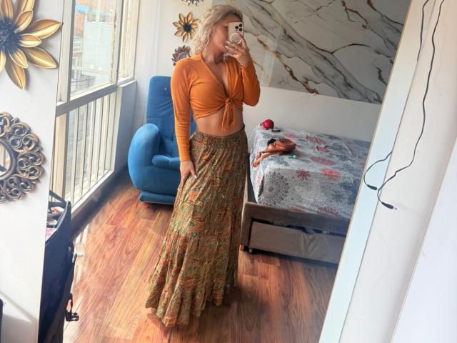 ElenaSaenz with curly blonde hair, orange crop top, and floral skirt, showing her tits in a mirror selfie, standing on wooden floor, holding phone in front of her face.