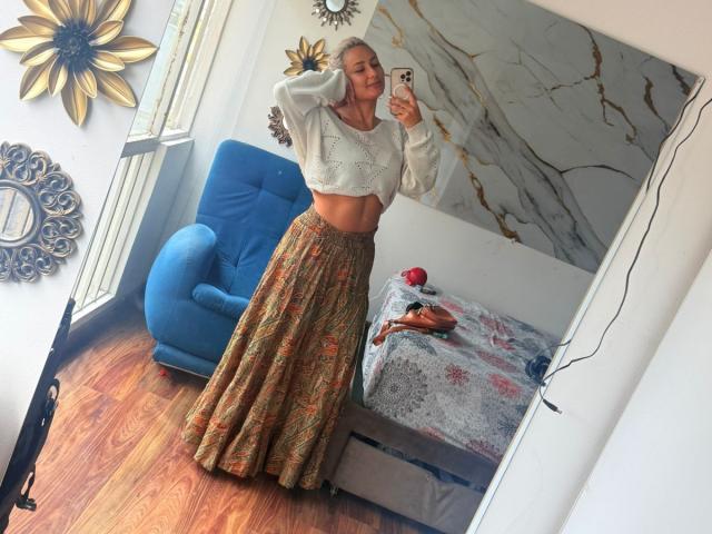 ElenaSaenz stands in a mirror selfie, wearing a white crop top and a long, colorful, floral skirt, with her tits slightly visible, holding a phone, smiling.