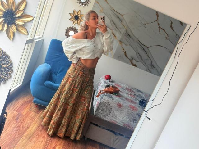 ElenaSaenz with a slim, toned midriff, wearing a white off-shoulder top, exposing small, perky tits, and a long, floral-patterned skirt, taking a mirror selfie.