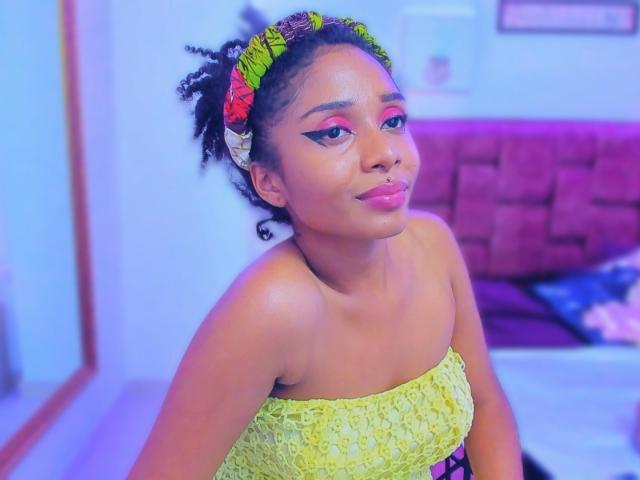 MeganWilliams with curly black hair in a yellow lace strapless dress, pink eyeliner, and neon headband, sitting with a cute smile, pink lipstick, and a sexy, confident look.