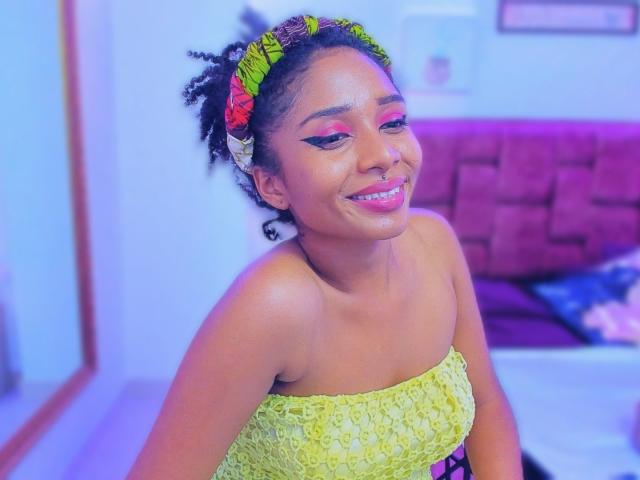 MeganWlliams with shiny brown skin, curly black hair in a neon green headband, pink eyeshadow, smiling with pink lipstick, wearing a yellow, textured strapless top, looking cute.