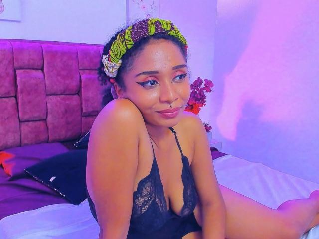 MeganWlliams with curly hair, green headband, black lace bra, sitting on bed, purple and pink lighting, resting chin on hand, cute, sexy, medium breasts, subtle smile, bedroom.