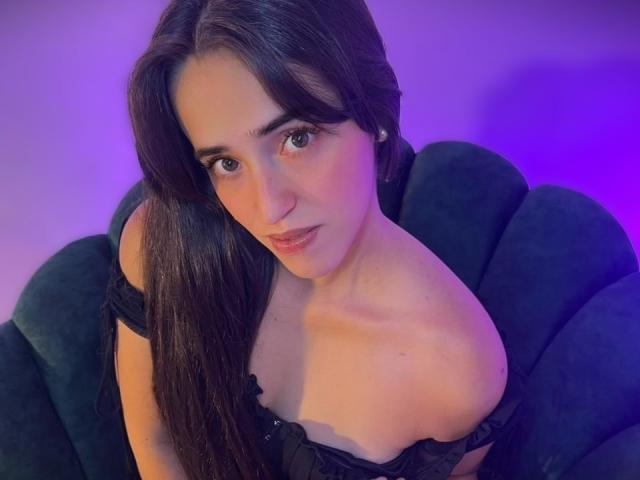 Camila Rossi, con lunghi capelli scuri, indossa una maglia nera con una spalla scoperta, che mette in mostra il seno e le spalle nude, seduta su una poltrona scura e imbottita, illuminata da luci viola e blu..