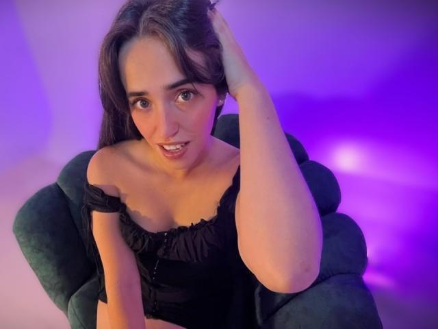 Camila Rossi, con cabello oscuro, vestida con una blusa negra con un hombro descubierto, sentada en una silla verde, con la mano en el cabello, sonriendo, iluminación morada y rosa, con los senos visibles, mirando hacia arriba..