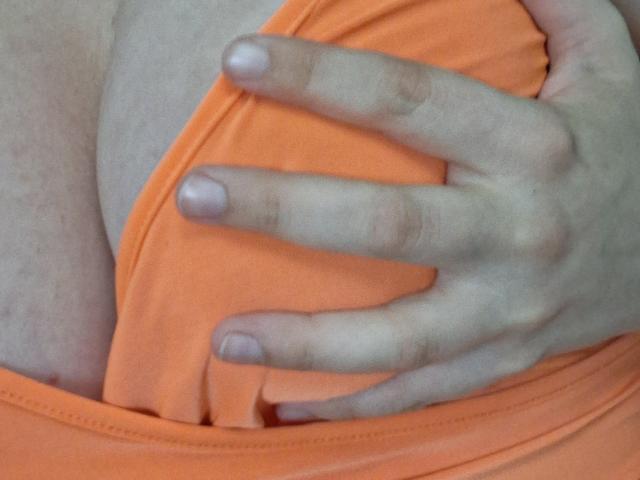 Marriam's hand grips her orange top, revealing her bare tits with pale skin and short, clean nails. Her fingers are slightly spread, showing her nipples just underneath.