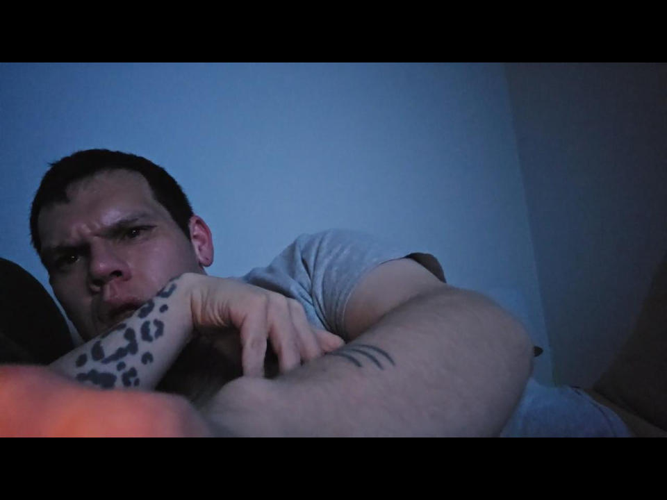 NightWolmy, a chubby dude with short dark hair, leans on a leopard-print arm, showing a tattoo on his bicep, with a slightly annoyed expression, in dim blue lighting.