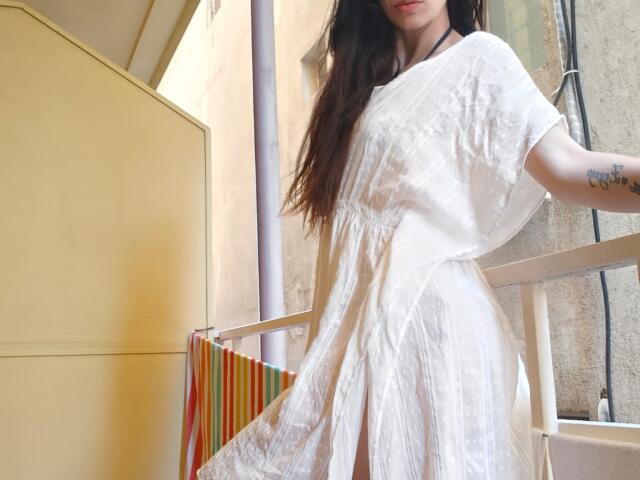 DanaWaw with long dark hair, wearing a white, semi-transparent dress, showing her tits and a tattoo on her right arm, standing by a balcony with colorful towels.