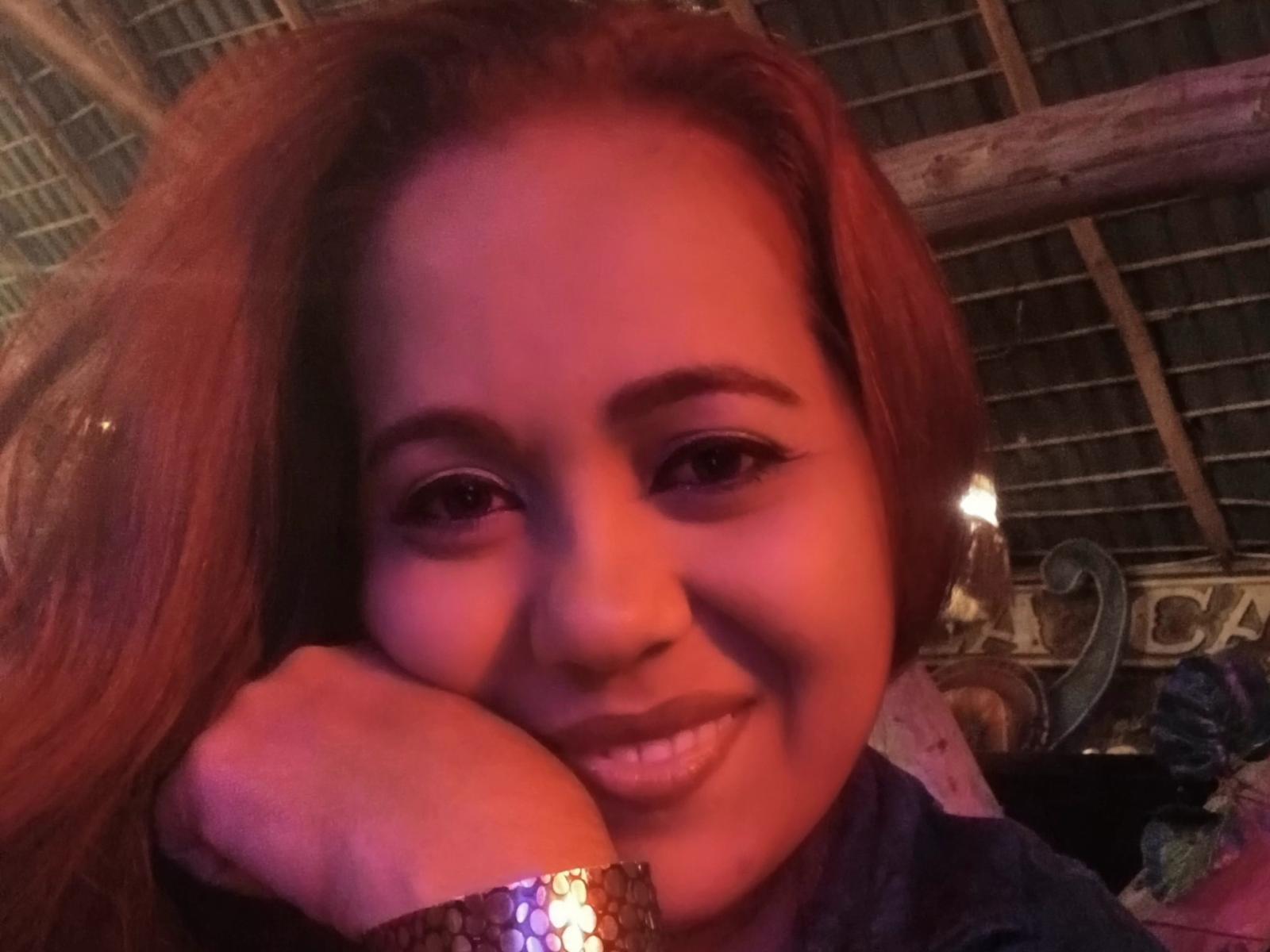 AfroditaTits with red hair, smiling, head resting on hand, wearing a metallic bracelet, soft pink-red lighting, wooden ceiling, and a small candle in the background.