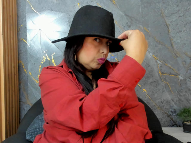 IvonnaLune with black hat, red shirt, and long black hair, pouting with purple lipstick, holding hat, showing her ass, sitting on black chair, in a room with marble wall.