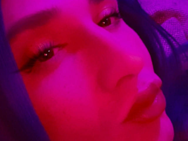 MaryXXL-hot's face with pink-purple lighting, full lips, long eyelashes, and dark hair, close-up, looking slightly to the side, showing subtle makeup and soft skin texture.