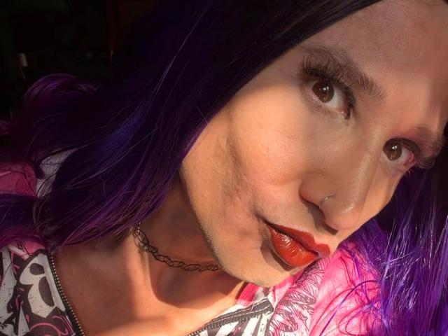 MaryXXL-hot with purple hair, red lipstick, nose ring, and choker, sunlight on her face, slight smile, showing her tits in a pink and black patterned top.