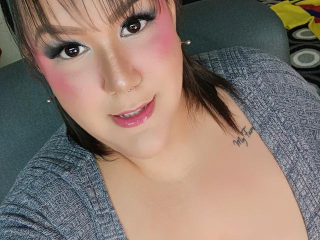 GiiannaMorrys with pink blush, black eyeliner, and nose ring, wearing a grey textured top, showing a tattoo on her chest, and a slight smile. Her tits are partially visible.