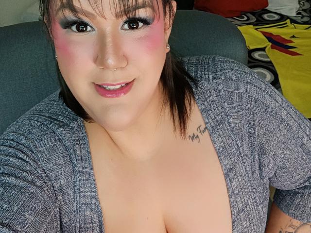 GiiannaMorrys with pink eyeshadow, nose ring, and small tattoo on chest, wearing gray patterned top, showing cleavage, smiling at camera. Her tits are slightly visible.