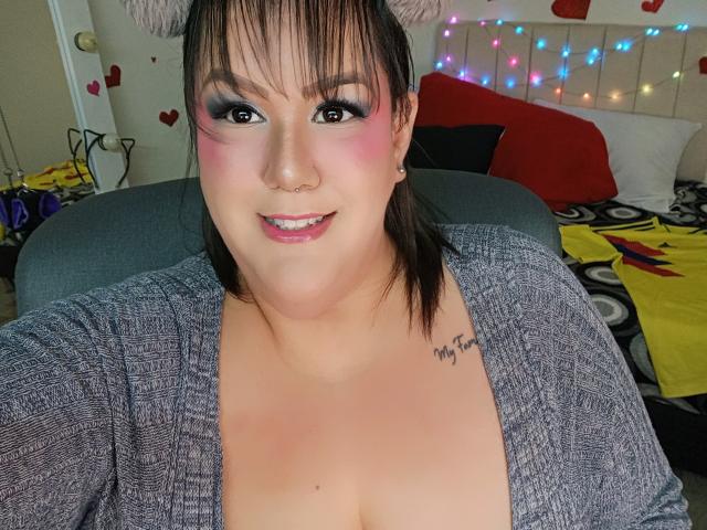 GiiannaMorrys, chubby with pink eyeshadow, grey cardigan, showing cleavage, small nose ring, tattoo on right shoulder, smiling, wearing furry cat ears, dark brown hair, pink lipstick.