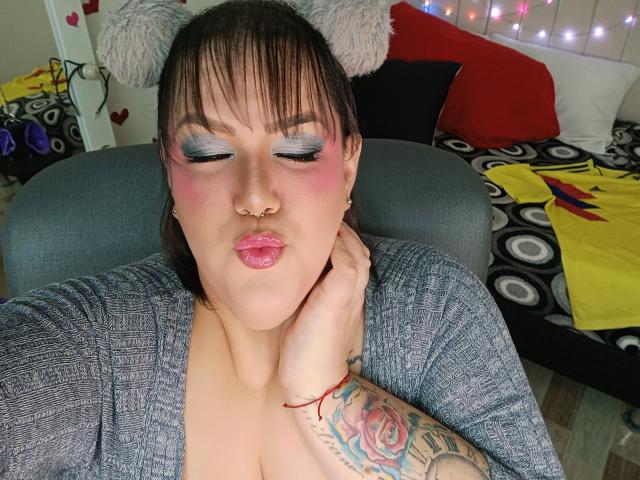 GiiannaMorrys with pink pout, blue eyeshadow, nose ring, gray cardigan, furry bear ears, tattooed arm, and puffy lips, eyes closed, hand on neck, sitting on gray chair.