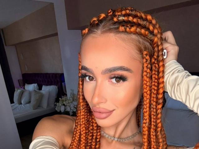 MarissaFex hat orangefarbenes Zopfhaar, große braune Augen und volle, rosafarbene Lippen. Sie trägt eine Kette und ein Oberteil mit einem Schulterteil, der offen ist, und hat eine Hand in ihrem Haar, während sie in die Kamera schaut..
