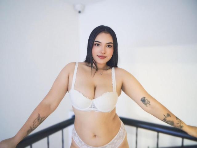 KendallCurves with medium-sized tits, wearing white lace bra and panties, standing with arms on black rail, tattoos on arms, black hair, light skin, slight smile, bedroom setting.
