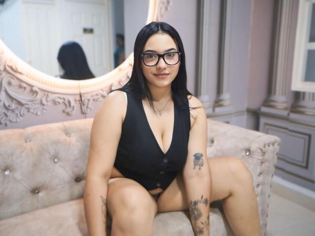 KendallCurves, con gafas, cabello negro y una blusa negra de escote bajo, mostrando sus pechos y tatuajes, sentada en un sofá beige con capitonado, sonriendo a la cámara..
