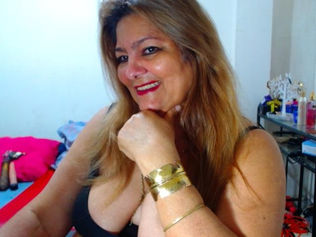 MujerCaliente69 con cabello rubio, lápiz labial rojo, blusa negra, mostrando senos grandes, pulseras doradas, sonriendo, sosteniendo su barbilla, en un dormitorio con almohadas rosas y rojas..