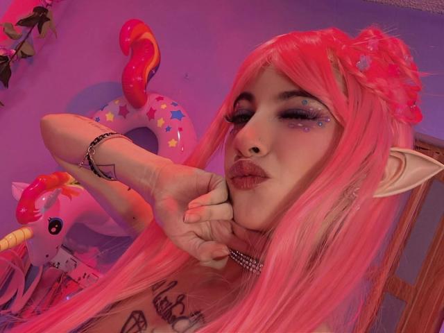 LaurenEvie69 with pink hair, elf ears, glittery eyeliner, nose ring, and choker, pouting with hand on chin, tattoo on chest, in pink-lit room with unicorn inflatable.