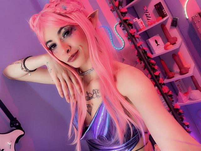 LaurenEvie69 with pink hair, elf ears, blue eyeliner, and a shiny purple top, showing her tits and a tattoo, leaning on a wall with a seductive smile.