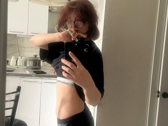 SophiaRenlay, avec des lunettes, des cheveux bruns ondulés, un t-shirt noir et des sous-vêtements noirs, exposant ses fesses fines et plates, et ses petites seins, tenant un téléphone pour un selfie devant un miroir..