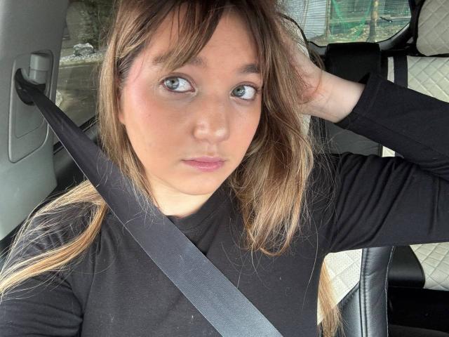 Michiko Vivona, com cabelo castanho, vestindo uma camisa preta, com o cinto de segurança atravessado no peito, com a mão no cabelo, olhando para o lado, olhos azuis, lábios rosados suaves, sentada em um carro..