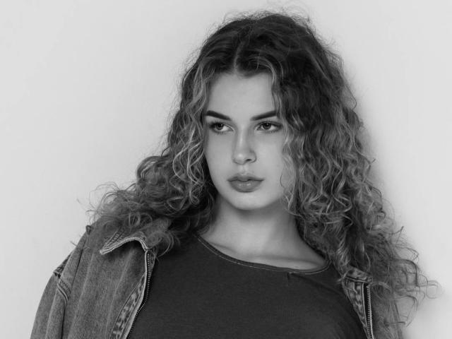LiaWanat, met lang krullend haar, draagt een denimjasje over een donkere blouse. Ze heeft een serieuze uitdrukking en haar borsten zijn gedeeltelijk zichtbaar onder de blouse. Zwart-wit foto..