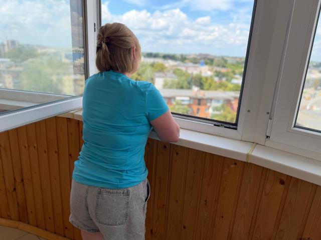 KiraSmiling with butt in gray shorts, blue shirt, and brown hair in a bun, leaning on a window, looking outside, wooden wall, white window frame, blue sky.