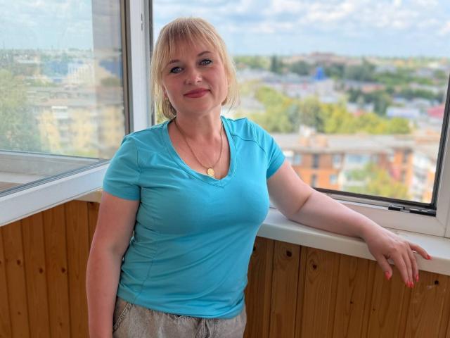KiraSmiling has blonde hair, blue eyes, and is wearing a tight blue shirt that shows her small tits and a gold necklace. She's leaning on a window with wood paneling.