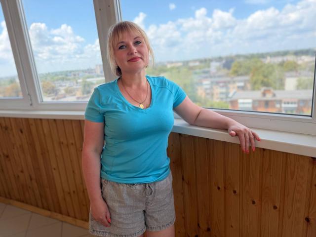 KiraSmiling, blonde woman with medium tits, wearing a blue top and gray shorts, leans on a wooden window ledge, smiling, with a bright blue sky outside.